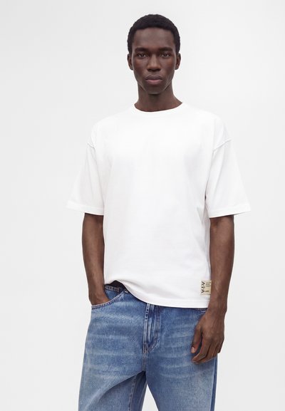 Homme à la peau foncée portant un t-shirt blanc uni à manches courtes et un jean bleu en denim, debout devant un fond clair uni.