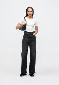 Μαύροι τζιν με ψηλή μέση και φαρδύ πόδι, συνδυασμένοι με εφαρμοστό λευκό t-shirt και μπλε τσάντα. Το μοντέλο φορά γυαλιστερά μαύρα παπούτσια και έχει πλεξούδες στα μαλλιά.