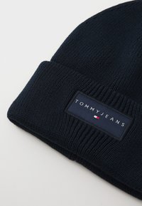 Navy knit beanie with a turned-up cuff and a rectangular fabric tag featuring the brand name "TOMMYJEANS" and a small logo.