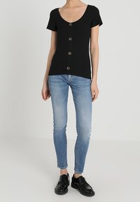 Top corto de manga corta negro con un escote redondeado y cinco botones marrones en la parte delantera, combinado con jeans ajustados azul claro y zapatos negros.