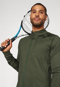 Grønn zip-up treningsjakke med høy krage og sidelomme, båret av en person som holder en tennisracket med blå aksent.