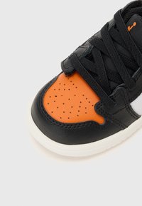 Black and orange sneaker featuring a smooth leather upper, perforated toe box, and black laces, with a white rubber sole and stitching details.