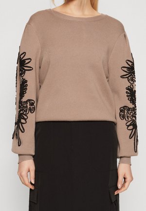 Pull beige avec broderie florale noire sur les manches, porté avec une jupe noire dotée de grandes poches avant.