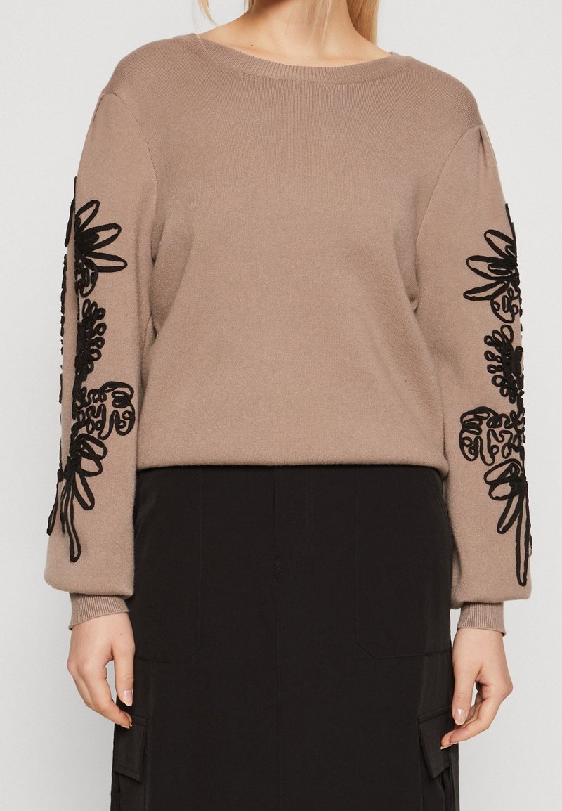Pull beige avec broderie florale noire sur les manches, porté avec une jupe noire dotée de grandes poches avant.
