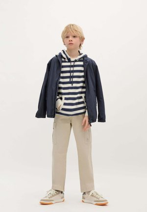 Giacca leggera navy sopra una felpa con cappuccio a strisce bianche e navy, pantaloni cargo beige e sneakers beige con lacci bianchi.