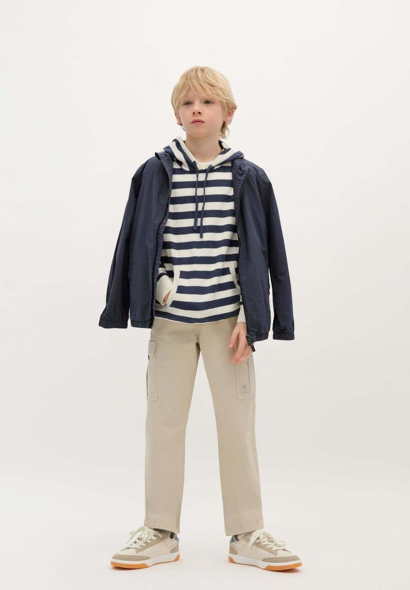 Marineblaue leichte Jacke über einem weißen und marineblau gestreiften Kapuzenpullover, beigen Cargo-Hosen und beigen Turnschuhen mit weißen Schnürsenkeln.