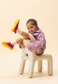 Ein weiß gepunkteter Stuhl mit abgerundeten Kanten hält ein Kind, das eine helllila Jacke, beigefarbene Hosen und farbenfrohe gelbe und rosa Schuhe trägt.