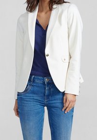 Vitt kavaj med skräddarsydd passform, enkel knäppning och guldaccent. Bärs över en marinblå topp, i kombination med blå jeans. Slät tyg.