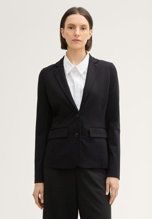 Schwarze taillierte Blazerjacke mit langen Ärmeln, zwei frontalen Taschen und Knopfverschluss. Kombiniert mit einem weißen Hemd mit Kragen darunter.