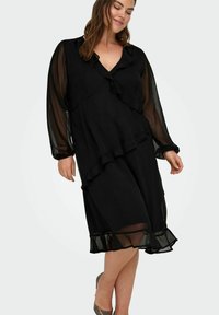 Czarna sukienka z falbanami, długimi przezroczystymi rękawami, dekoltem w kształcie litery V i warstwową spódnicą. Gładka tekstura, długość do kolan.