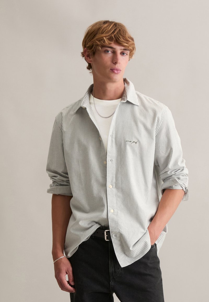 Junger Mann mit blonden Haaren, der ein hellgraues gestreiftes Hemd über einem weißen T-Shirt, schwarze Hosen, eine silberne Kettenhalskette und ein Armband trägt, vor schlichtem Hintergrund steht.