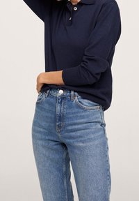 Granatowy sweter typu polo z dzianiny z kołnierzykiem, zestawiony z wysokim stanem jasnoniebieskich dżinsów. Szczegóły obejmują zapięcie na guziki i boczne kieszenie.