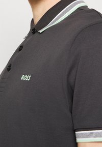Polo en charbon avec un tissu texturé, trois boutons, et un col rayé avec des accents vert menthe et un logo sur la poitrine.