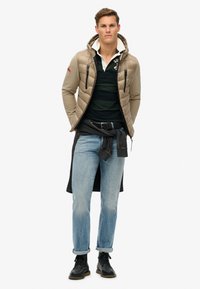 Lettene, beige dunjakke med hætte, lynlåslommer; sort og grøn stribet polo skjorte; blå jeans; sorte støvler.