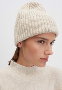Beanie lavorato a maglia di colore crema con coste verticali e una parte superiore leggermente conica, abbinato a un maglione beige con collo alto.