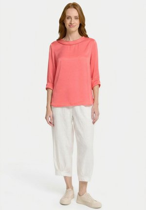 Woman with shoulder-length hair wearing a coral long-sleeve top, white cropped pants, and beige sneakers, standing against a white background.