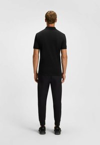 T-shirt polo noir à manches courtes, en tissu texturé, avec un col, associé à un pantalon noir fuselé et des baskets noires, vu de dos.