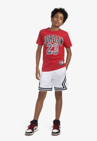 Maglietta rossa a maniche corte con la scritta "JORDAN 23" in grande carattere nero. Pantaloncini da basket bianchi con dettagli neri e strisce. Scarpe da ginnastica nere e rosse.