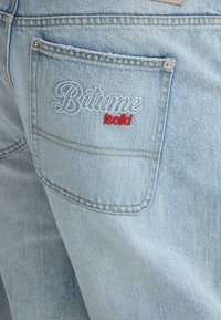 Jean en denim bleu clair avec une poche arrière ornée d'un texte brodé en blanc et rouge. Texture lisse avec des détails de couture standard.