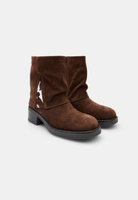 Stivali alla caviglia in suede marrone con un design morbido, caratterizzati da un taglio diagonale e un tacco a blocco. Texture morbida e dettagli di cuciture a contrasto.