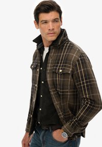 Camisa de cuadros en tonos marrones y verdes con cuello, detalles de bolsillos y botones a presión, llevada sobre una camiseta oscura y jeans azules.