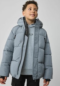 Doudoune grise avec un design matelassé, une capuche et des éléments en métal noir contrastants. Présente une marque sur la fermeture éclair et les sangles.