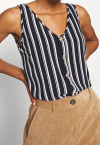 Femme portant un haut à rayures verticales noires et blanches sans manches, rentré dans un pantalon en velours côtelé beige taille haute avec une fermeture à boutons.