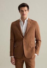 Giacca di un completo in lino marrone, con rever a contorno, chiusura a due bottoni e tasche laterali, abbinata a una camicia bianca sotto.