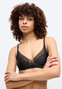 Γυναίκα με σγουρά μαλλιά που φοράει μαύρο σουτιέν με δαντέλα, με σταυρωμένα τα χέρια, μπροστά σε ένα απλό ανοιχτό φόντο.