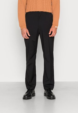 Mann, der schwarze maßgeschneiderte Hose, schwarze Leder-Schnürschuhe und einen braunen, grob gestrickten Pullover trägt, steht auf einem grauen Boden.