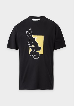Mand iført sort T-shirt med stor hvid tegneserie-kanin og gul firkant, mørkeblå jeans og sorte sko, stående med hænderne i lommerne.