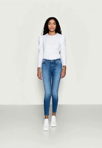 Jonge vrouw met krullend haar, gekleed in een witte blouse met lange mouwen, blauwe skinny jeans en witte sneakers, staand voor een effen achtergrond.