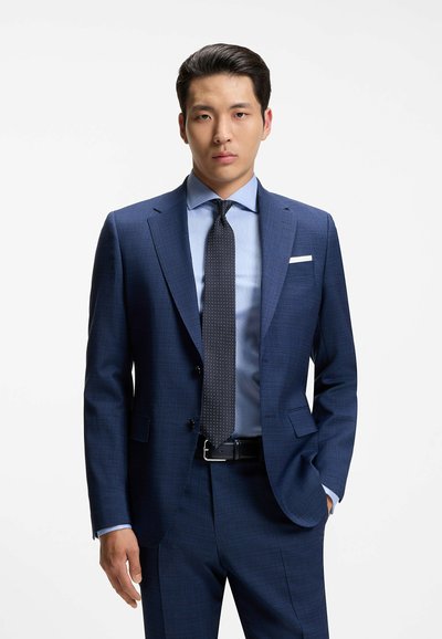 Joven con un traje azul a cuadros, camisa azul clara y corbata negra con puntos, de pie con una mano en el bolsillo contra un fondo blanco.