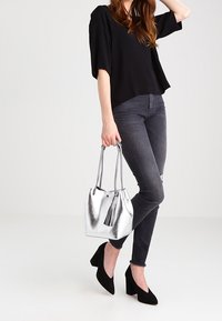 Sac en toile argent métallique avec un pompon, associé à un haut noir court et un jean skinny gris foncé, porté avec des chaussures à talons hauts noires.