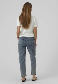 Camicia bianca a maniche corte, jeans dritti blu chiaro e sandali neri. Capelli lunghi e lisci castani scendono oltre le spalle.