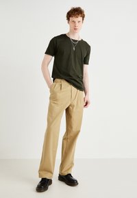 Olive green t-shirt, beige trousers with a loose fit, and black shoes. Model wears layered silver necklaces. Simple, casual attire.