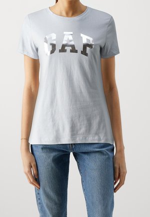 Woman wearing light blue fitted short-sleeve T-shirt with shiny silver "GAP" logo paired with blue denim jeans, standing against a neutral background.