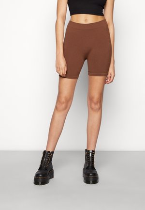 Frau trägt braune gerippte Biker-Shorts, ein schwarzes Crop-Top und schwarze Schnür-Plateau-Stiefel, steht an einer weißen Wand.
