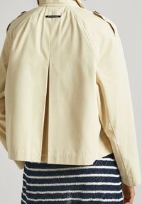 Chaqueta corta de un beige claro, hecha de un tejido suave, con un pliegue en la parte trasera y epauletes en los hombros. Abajo, una falda de rayas con textura.