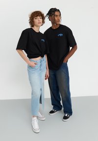 Juodos medvilnės marškinėliai su spalvingu logotipu, šviesiai mėlyna džinsinė sijonas su šonine įpjova ir tamsūs džinsai. Modelis dėvi baltus ir juodus sportinius batelius.
