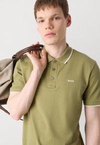 Olivno zelena polo majica s ovratnikom, svijetlosmeđim detaljima, tri gumba i BOSS logotipom s lijeve strane. Model drži torbu na ramenu.