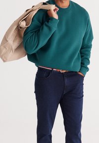 Bluza w kolorze morskim z okrągłym dekoltem, zestawiona z ciemnoniebieskimi dżinsami. Beżowa torba na ramię przewieszona przez ramię. Prosty fason, teksturowana tkanina.