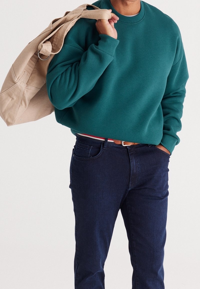 Bluza w kolorze morskim z okrągłym dekoltem, zestawiona z ciemnoniebieskimi dżinsami. Beżowa torba na ramię przewieszona przez ramię. Prosty fason, teksturowana tkanina.