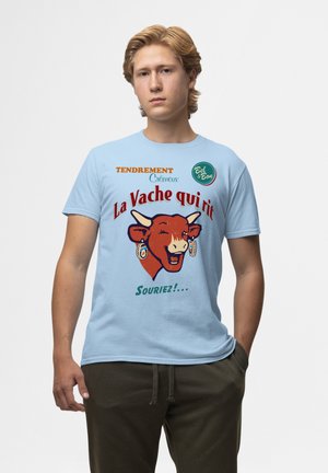 Světle modré tričko s grafikou usmívající se krávy s náušnicemi, textem "La Vache qui rit" a zelenými barevnými akcenty. Pohodlný střih.