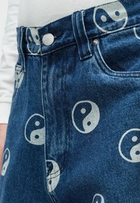 Modri denim kavbojke z belimi natisnjenimi yin-yang simboli, srebrno zaponko in delno vidna bela majica s dolgimi rokavi.