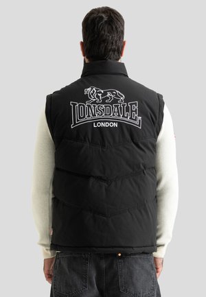 Homme portant une veste sans manches matelassée noire avec le logo Lonsdale London et un motif de lion dans le dos, une chemise blanche à manches longues et un jean foncé.