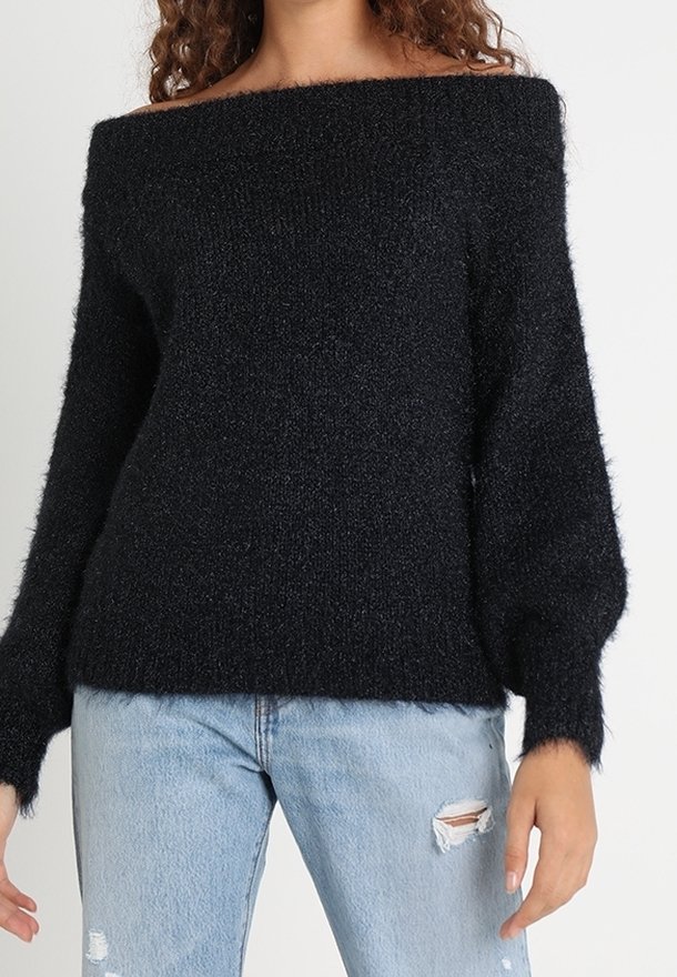 Femme portant un pull noir à épaules dénudées en matière pelucheuse avec manches longues et un jean bleu clair déchiré sur un fond uni.