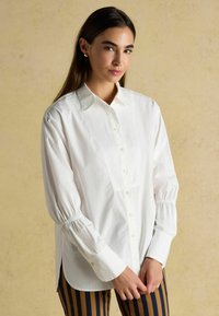 Weißes Hemd mit strukturiertem Kragen, gerafften Ärmeln und Seitenschlitzen. Getragen mit marineblauen und beigen gestreiften Hosen. Glatte Textur.