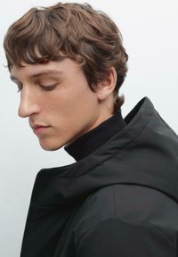 Schwarze Jacke mit hohem Kragen, die über einem schwarzen Rollkragenpullover getragen wird. Der Stoff wirkt glatt mit matter Oberfläche. Haare in sanften Wellen gestylt.