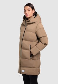 Abrigo de plumas largo en color beige con capucha, que cuenta con una cremallera frontal, dos bolsillos laterales y detalles en negro. Textura suave y diseño acolchado.